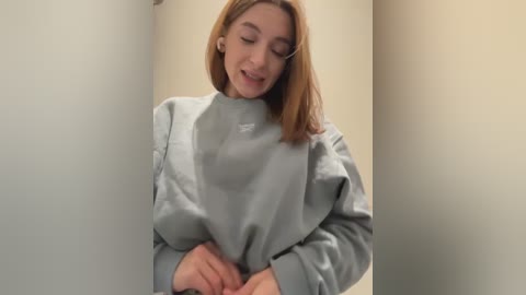 Media: Video of a young Caucasian woman with shoulder-length auburn hair, wearing a light blue medical gown, smiling while adjusting her sleeves, in a sterile hospital corridor.