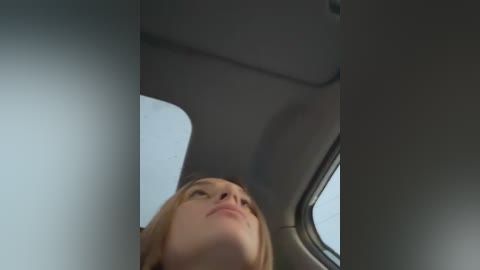 Media: A video of a blonde woman with fair skin, looking upwards, inside a car. The image is slightly out of focus, capturing the roof and window of the vehicle.