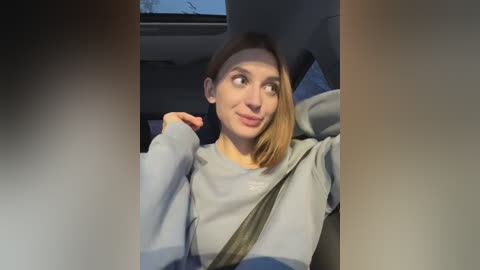 Media: Video of a smiling young woman with shoulder-length blonde hair, wearing a gray sweatshirt, driving a car with a gray seatbelt across her chest, window with tree branches visible in the background.