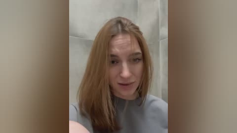 Media: A video of a fair-skinned woman with straight, shoulder-length brown hair, wearing a grey top, slightly smiling, in a blurred bathroom setting with beige tiles.