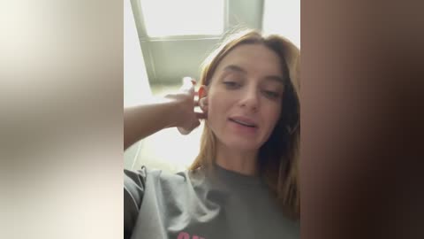 Media: Video of a young Caucasian woman with light brown hair and fair skin, wearing a gray T-shirt, being examined by a doctor in a clinical setting with a light box overhead.