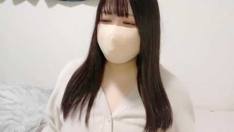 Media: Video of an Asian woman with long, straight black hair, wearing a beige face mask, white cardigan, and a white top. She has a fair complexion and is standing against a plain white wall with a gray blanket behind her.