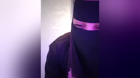 Media: Video of a person wearing a black mask, partially visible, standing against a white wall under purple lighting. The person's face is obscured, creating a sense of mystery and intrigue.