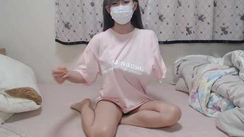 Media: Video of an Asian woman with fair skin, long black hair, and a white surgical mask, wearing a loose pink t-shirt, sitting on a bed with a floral-patterned bedspread, and a white blanket nearby.