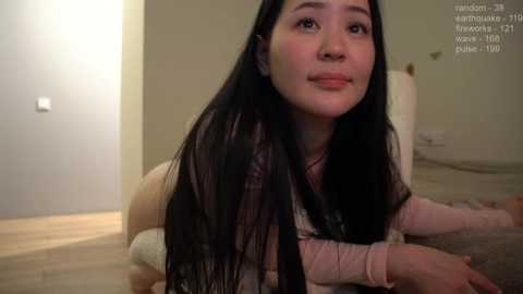 Media: Video of an Asian woman with long black hair, wearing a pink shirt, lying on her stomach on a beige rug in a minimalist, beige-toned room.