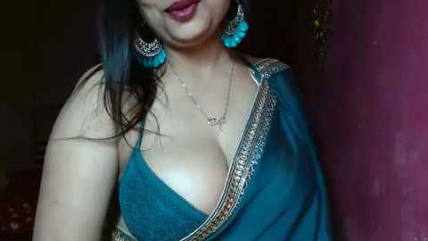 Media: Video of a South Asian woman with medium skin tone, wearing a teal sari with gold embroidery, revealing cleavage, and turquoise earrings. She has long, straight black hair and is smiling.