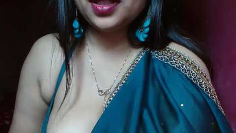 Media: Video of a South Asian woman with fair skin, wearing a teal sari with gold embroidery, turquoise flower earrings, and a silver necklace, smiling.