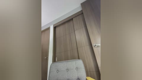 Media: Video of a modern, minimalist room with a light brown wooden wardrobe and sliding door, a gray upholstered chair with button tufting, and a yellow pillow on the floor.