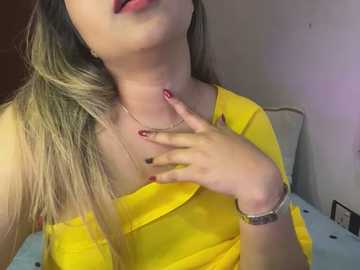 Media: A video of a woman with light skin and long, blonde hair, wearing a yellow off-shoulder top and a silver necklace, touching her chest, against a plain white wall background.