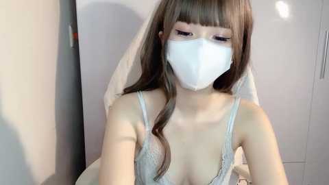 Media: A video of an Asian woman with long, straight brown hair, wearing a white face mask and a sheer, white lace camisole, seated indoors with a white wall and door in the background.