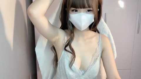 Media: Video of an East Asian woman with long, wavy brown hair, wearing a white face mask, light blue lace bra, and a white towel draped over her shoulder, posing in a white-lit room.