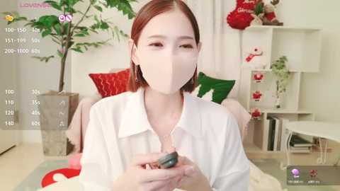 Media: Video of an East Asian woman with straight, shoulder-length brown hair, wearing a white blouse, a face mask, and holding a green object, indoors with a white background, green plant, and red decorations.