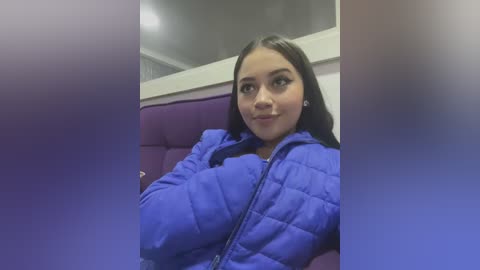 Media: A video of a young woman with straight, dark hair, wearing a blue puffer jacket, seated in a purple cushioned chair in a dimly lit room with a white ceiling.