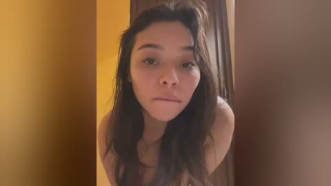 Media: Video of a young woman with long, dark hair, light skin, and a neutral expression, leaning against a wooden door in a warmly lit room with mustard-yellow walls.