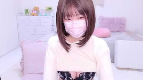 Media: Video of a young East Asian woman with straight brown hair, wearing a white long-sleeve shirt and black lace bra, face mask, and pink gloves, in a pastel-colored bedroom with white furniture and floral decor.