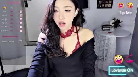 Media: A video of a young Asian woman with long black hair, wearing a black shirt and red lace bra, in a modern, minimalist room with a white fridge and table.