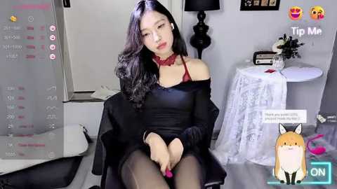 Media: Video of an East Asian woman with long black hair, wearing a black off-shoulder dress, sitting on a white chair in a modern room with a TV screen showing a live stream.