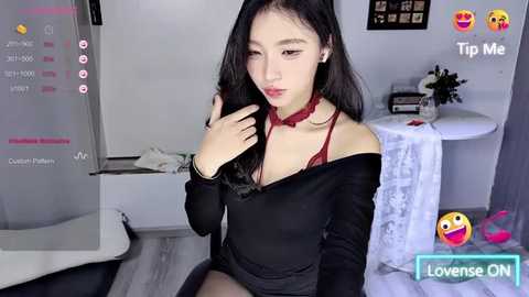 Media: A young Asian woman with long black hair and fair skin, wearing a black off-shoulder top and red lace choker, sits on a white chair in a modern, minimalistic room.