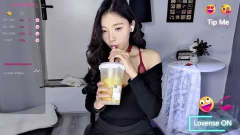 Media: Video of an Asian woman with long black hair, wearing a black off-shoulder top and black pants, drinking a clear drink. Background features a white wall, a small table, and a TV showing \"Lowrise ON\" with emojis.