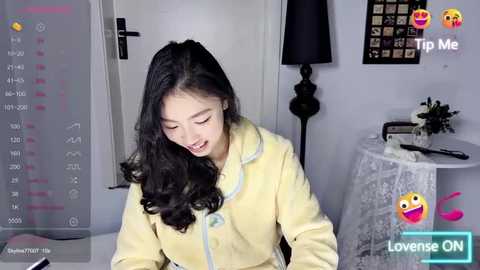 Media: A video of an Asian woman with long black hair, wearing a yellow jacket, smiling at a laptop, in a modern, minimalistic bedroom.