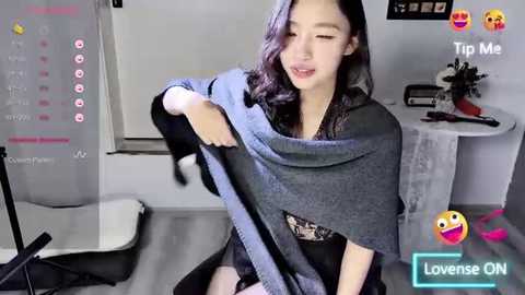 Media: Video of an Asian woman with long, wavy black hair, wearing a grey cardigan, black lace top, and black skirt, sitting in a modern office setting.