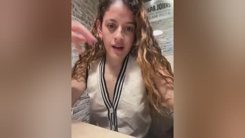 Media: A candid video of a young woman with long, wavy brown hair, wearing a white, sleeveless top with black stripes, in a pizza restaurant with exposed brick walls.