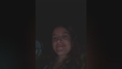 Media: A video of a young woman with curly dark hair, wearing a black jacket, smiling, dimly lit, and framed by a dark background.