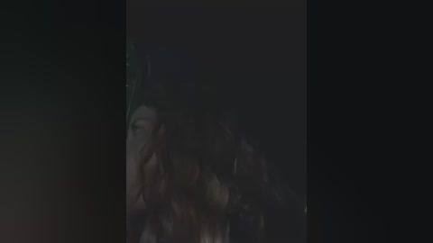 Media: A dimly lit video of a young woman with long dark hair, partially obscured by shadows, holding a phone close to her face, suggesting a private moment. The background is dark and indistinct.