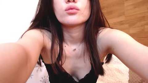 Media: A video of a young East Asian woman with long, straight black hair, fair skin, and a slender build, wearing a black lace bra, puckering her lips in a kiss. Background features a wooden wall and beige carpet.