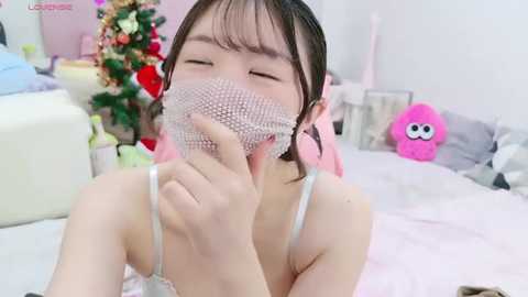 Media: Video of an Asian woman with light skin and brown hair, wearing a white lace mask, pink bow, and a pink tank top, in a bright, cluttered room with a Christmas tree and plush toys.