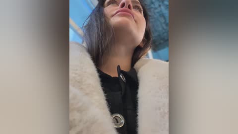 Media: A video of a woman with fair skin, wearing glasses, a black top, and a white fur-trimmed coat, standing outdoors with a blue sky and blurred background.