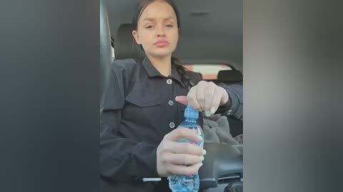 Media: Video of a woman in a car, wearing a black jacket, drinking water from a plastic bottle. She has long dark hair and a neutral expression.