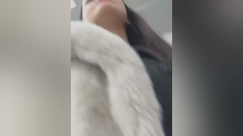 Media: Video of a close-up, blurred face with long dark hair, partially covered by a white fur-trimmed coat. The background is out of focus, giving a soft, intimate feel.