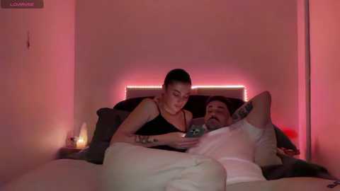 Media: Video of a woman with short hair, black tank top, and white pajama bottoms, lying in bed with a man, watching a tablet.