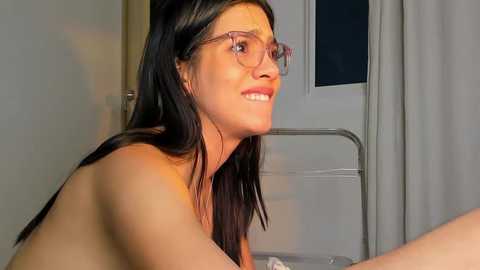 Media: Video of a topless, fair-skinned woman with long, straight black hair, wearing glasses, smiling. She's in a hospital room with white walls and a metal bed frame in the background.