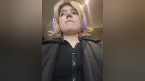 Media: Video of a woman with fair skin, shoulder-length blonde hair with pastel pink and purple streaks, wearing a black hoodie and purple headphones. She gazes upwards, looking thoughtful.