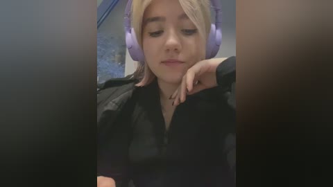 Media: A video of a young woman with light skin and blonde hair, wearing large purple headphones and a black shirt, leaning on a table with a neutral background.