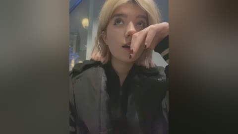 Media: A video of a young woman with platinum blonde hair, fair skin, and a neutral expression, wearing a black jacket, her fingers near her mouth, taken indoors with blurred background.