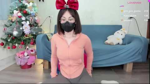 Media: Video of an Asian woman with straight, shoulder-length brown hair, wearing a black face mask, red bow headband, and peach hoodie. She kneels in front of a blue couch with a plush white dog. A decorated Christmas tree is in the background.