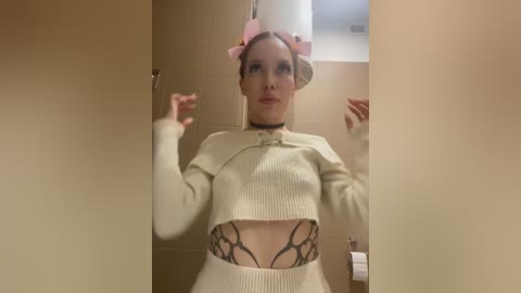 Media: Video of a light-skinned woman with a tattooed stomach, wearing a cream ribbed crop top and pink bow headband, standing in a beige-tiled bathroom.