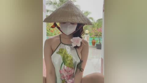 Media: Video of a young Asian woman with a straw hat, floral mask, and floral dress, sitting in an outdoor garden with tropical plants and colorful flowers in the background.