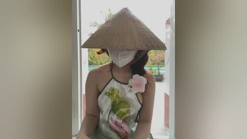 Media: A video of a woman with a conical hat, floral dress, and face mask, standing in a doorway with a garden view.