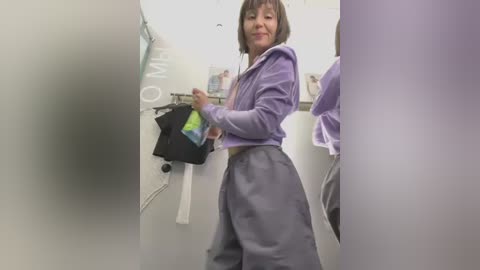 Media: Video of an Asian woman in a lavender hoodie and gray sweatpants, standing in a bathroom, holding a black bag with green and yellow patterns, surrounded by white stalls and a white sink.