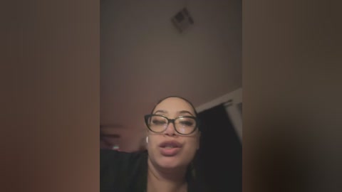 A video of a woman with light skin and dark hair, wearing glasses, making a kissy face. The background shows a dimly lit room with a ceiling light fixture and a partially visible curtain.