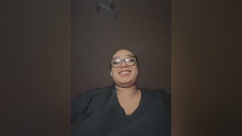 Media: Video of a smiling, bald woman with large round glasses, wearing a black shirt, against a dark, dimly lit background.