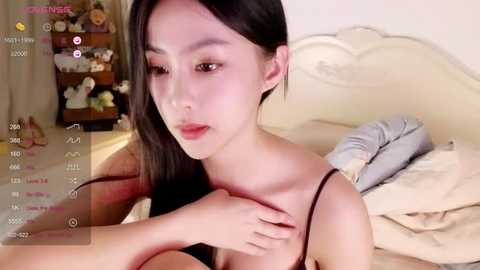 Media: Video of an East Asian woman with long black hair, fair skin, and a petite frame, wearing a black spaghetti strap top, sitting on a beige bed, surrounded by stuffed animals and a wooden shelf in the background.