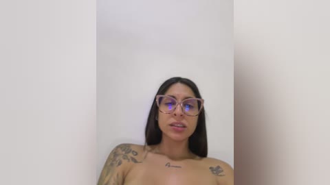 Video of a topless, light-skinned woman with long black hair, wearing glasses, tattoos on her arms, and sitting against a plain white wall.