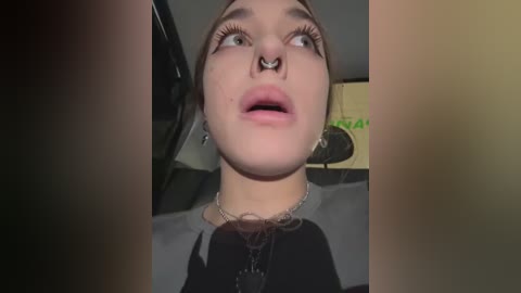 Media: Video of a young woman with light skin, wearing a septum ring, hoop earrings, and a black shirt. She has long eyelashes and is looking upward. Background shows a car interior and a green sign.