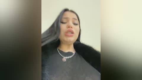 Media: A blurred video of a young woman with long black hair, wearing a black fur coat and silver necklace, standing against a plain, light-colored background.