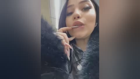 Media: Video of a Latina woman with long black hair, wearing black fur-trimmed jacket, seductively biting her finger, eyes half-closed, in a dimly lit, blurred background.